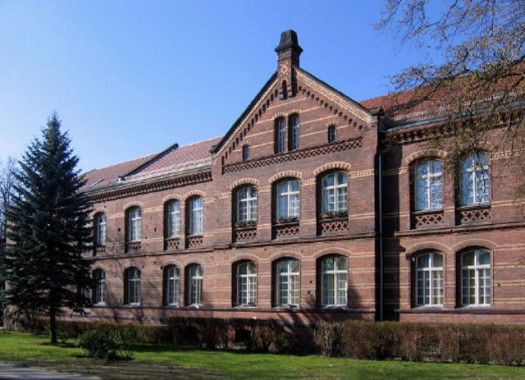 Ein zweistöckiges Backsteingebäude mit Bogenfenstern und Satteldach steht neben einem Baum und einer grünen Wiese unter einem klaren blauen Himmel.
