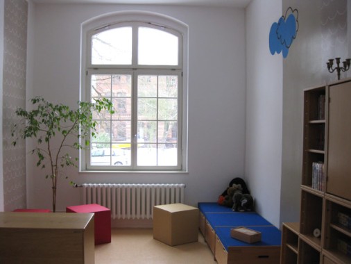 Ein helles, minimalistisches Spielzimmer mit einem großen Fenster, einer Blattpflanze, niedrigen Schränken, einer blauen gepolsterten Bank, Plüschtieren, Regalen und einer blauen Wolkendekoration an der Wand.