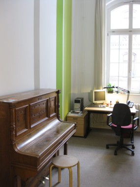 Ein hölzernes Klavier und ein Hocker stehen neben einem Schreibtisch mit Computer, Lampe und Stuhl in einem hellen Raum mit großen Fenstern und blassgrünen Akzenten.