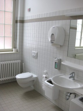 Eine saubere, moderne öffentliche Toilette mit einer Wandtoilette, einem Waschbecken, einem Spiegel, einem Seifenspender, einem Papierhandtuchspender und einem Fenster.