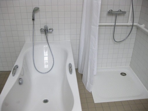 Ein Badezimmer mit einer weißen Badewanne und einer separaten Dusche, beide mit Handbrause, umgeben von weiß gefliesten Wänden.