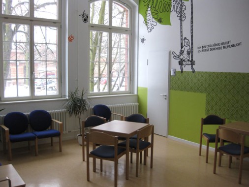 Ein Raum mit großen Fenstern, grünen und weißen Wänden, mehreren Holztischen und -stühlen, einer Topfpflanze und Wandkunst an der Decke.
