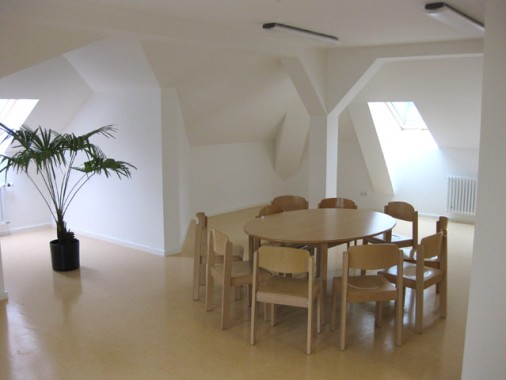 Ein runder Holztisch mit sieben Stühlen steht in einem hellen, spärlich eingerichteten Raum mit weißen Wänden, schrägen Decken und einer Topfpflanze.