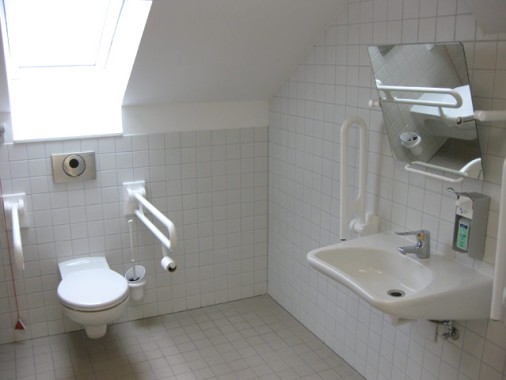 Barrierefreies Badezimmer mit weißen Fliesen, einer an der Wand befestigten Toilette und einem Waschbecken, Handläufen, einem kippbaren Spiegel, einem Papierhandtuchspender und einem Fenster, das für Tageslicht sorgt.