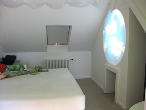 Ein minimalistisches Schlafzimmer im Dachgeschoss mit einem niedrigen Bett, einem Dachfenster und einer runden Projektion eines Wolkenhimmels an der Wand. Einige Spielzeuge oder Stofftiere liegen auf dem Bett.