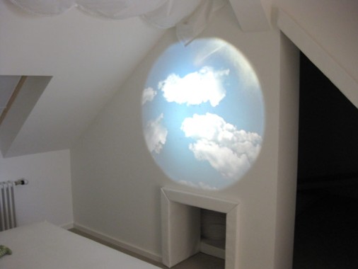 Eine Projektion eines blauen Himmels mit weißen Wolken wird auf eine weiße Wand in einem minimalistischen Dachgeschossraum projiziert.