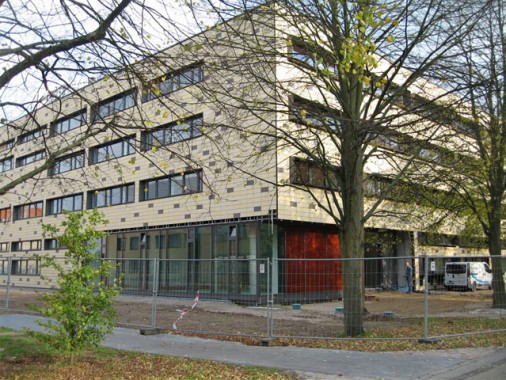 Ein modernes, vierstöckiges Gebäude im Bau, umgeben von einem Metallzaun, mit kahlen Bäumen und Baumaterialien im Vordergrund.