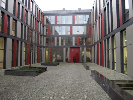 Moderner Innenhof mit Kopfsteinpflaster, rechteckigen Pflanzgefäßen und Glaswänden mit roten und grauen vertikalen Akzenten. Zwei Personen stehen in der Nähe des Eingangs am anderen Ende.