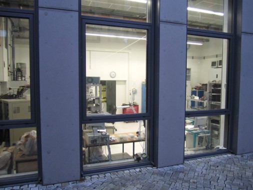 Blick durch große Fenster in ein gut beleuchtetes Labor, in dem wissenschaftliche Geräte, Maschinen und Arbeitsplätze zu sehen sind; außen mit Steinpflasterung.