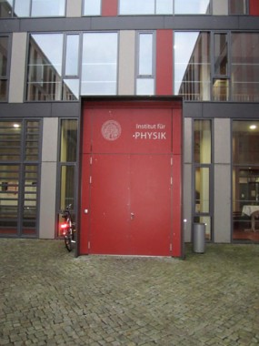 Eine rote Tür mit der Aufschrift Institut für Physik an einer modernen Gebäudefassade mit einem in der Nähe geparkten Fahrrad und einem Steinpflaster davor.