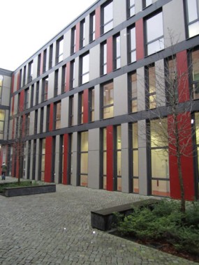 Ein modernes mehrstöckiges Gebäude mit vertikalen roten und grauen Paneelen, großen Fenstern und einem gepflasterten Innenhof mit Bänken und blattlosen Bäumen.