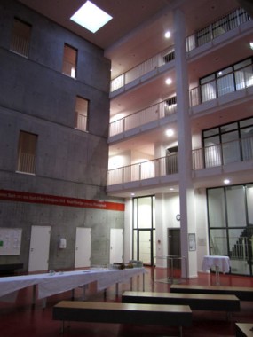 Ein modernes Innenatrium mit mehrstöckigen Balkonen, Betonwänden, rotem Bodenbelag, Tischen mit weißen Tischdecken und Bänken.