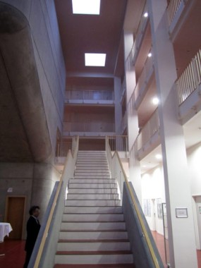Eine breite Treppe führt zu mehreren Stockwerken in einem modernen Gebäude mit weißen Geländern, Betonwänden und Oberlichtern; links von der Treppe steht eine Person.
