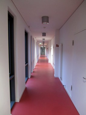 Ein langer, leerer Flur mit rotem Fußboden, weißen Wänden, mehreren Türen auf der rechten Seite und Fenstern auf der linken Seite, beleuchtet von Deckenlampen.
