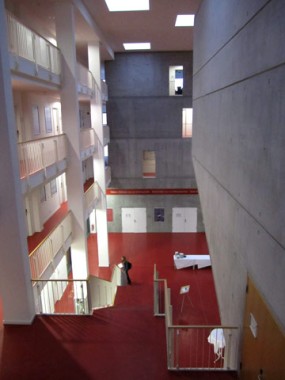 Eine Person steht auf einem roten Boden in einem modernen, mehrstöckigen Gebäude mit Sichtbetonwänden, weißen Geländern und Treppenhäusern.