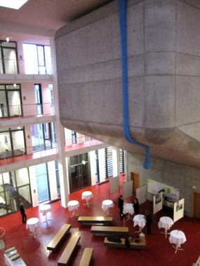 Modernes Interieur mit roten Böden, Betonwänden, einer großen Hängekonstruktion mit einer blauen Pfeife, runden Tischen mit weißen Tischdecken, Bänken und darunter stehenden Menschen.