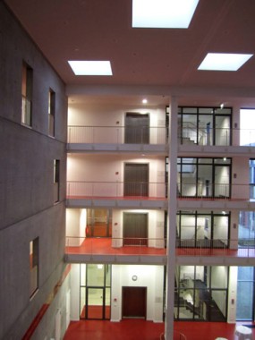 Innenansicht eines modernen mehrstöckigen Gebäudes mit roten Böden, Betonwänden, verglasten Räumen und großen Deckenleuchten.