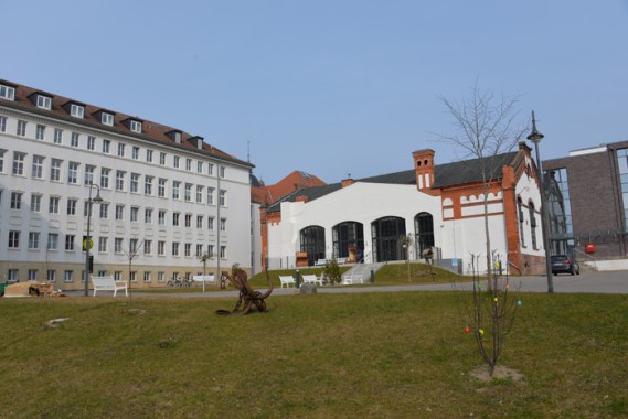 Eine Rasenfläche mit Bänken und kleinen Bäumen vor zwei großen Gebäuden, eines weiß und modern, das andere mit roten Backsteinakzenten und Bogenfenstern.