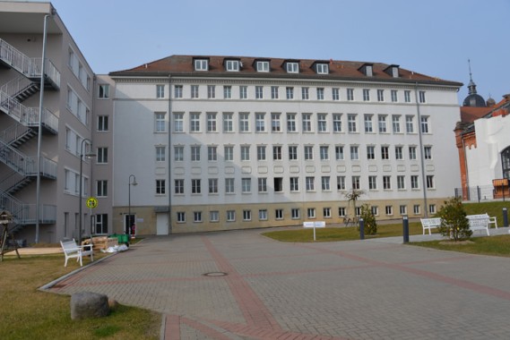 Ein großes weißes, mehrstöckiges Gebäude mit vielen Fenstern, das an einen gepflasterten Innenhof mit Bänken, etwas Gras und einer Metalltreppe auf der linken Seite angrenzt.