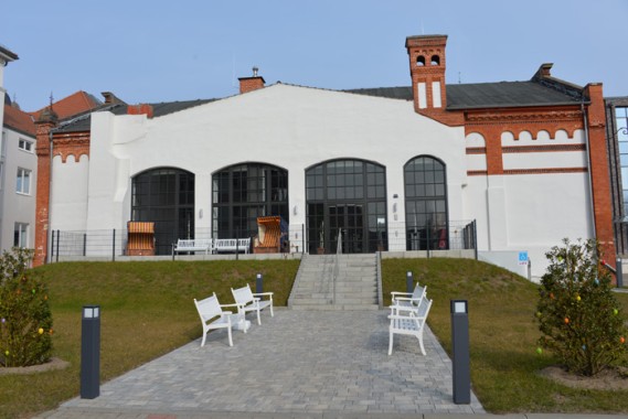 Ein weißes Backsteingebäude mit großen Bogenfenstern, Außenbänken und einem gepflasterten Weg, der zum Eingang führt, umgeben von Gras und kleinen Büschen.