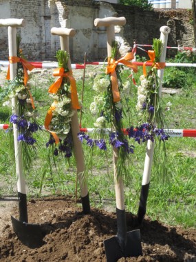 Drei Schaufeln mit Blumen und orangefarbenen Bändern stehen aufrecht in der Erde auf einer Baustelle, wahrscheinlich für einen ersten Spatenstich.