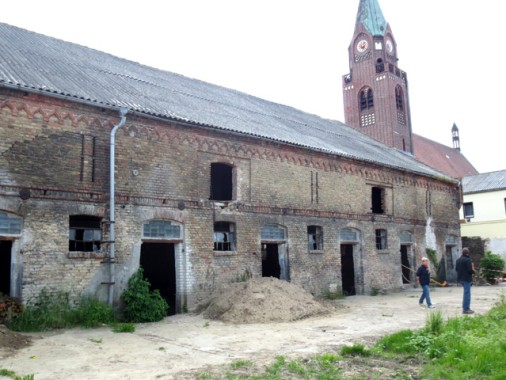 Ein langgestrecktes Backsteingebäude mit mehreren offenen Türen und Fenstern steht vor einem Kirchturm; zwei Personen betreten den Hof.