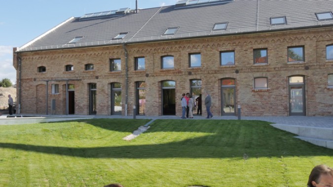 Eine Gruppe von Menschen steht vor einem renovierten Backsteingebäude mit großen Fenstern und Türen, umgeben von einem Rasen und gepflasterten Wegen.
