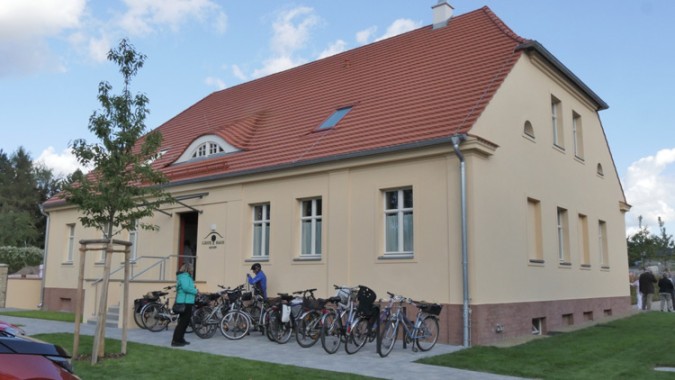 Ein beigefarbenes, zweistöckiges Gebäude mit rotem Ziegeldach, mehrere Fahrräder, die draußen geparkt sind, und ein paar Leute, die in der Nähe des Eingangs stehen.
