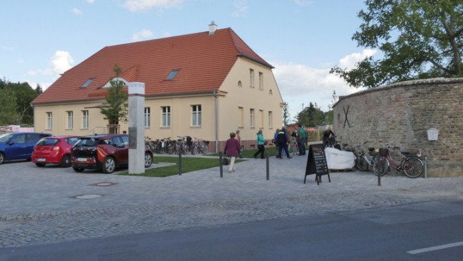 Ein beigefarbenes zweistöckiges Gebäude mit rotem Dach, mehrere geparkte Autos, Menschen, die sich neben Fahrrädern versammelt haben, und eine Steinmauer entlang einer gepflasterten Straße.