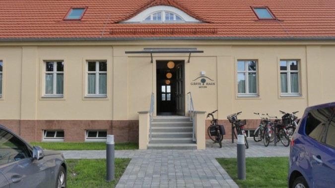 Vordereingang eines beigen Gästehauses mit orangefarbenem Ziegeldach, mehreren Fenstern, abgestellten Fahrrädern und zwei Autos, die teilweise auf beiden Seiten sichtbar sind.