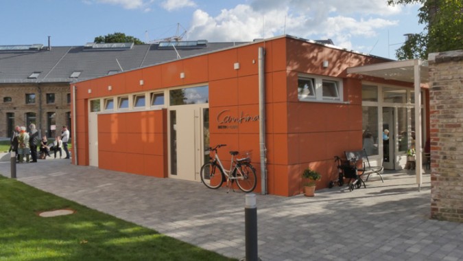 Einstöckiges, modernes, orangefarbenes Gebäude mit großen Fenstern, einem Schild mit der Aufschrift Cantina, zwei Fahrrädern, einer Topfpflanze und einer Menschenansammlung im Hintergrund.