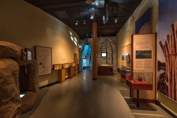 Eine Museumsausstellung mit Informationstafeln, Vitrinen mit Artefakten und einer zentralen Holzsäule, alles bei warmer Innenbeleuchtung.