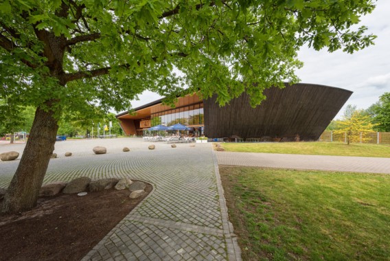 Modernes Gebäude mit abgewinkelter Holzfassade, umgeben von Bäumen und einem gepflasterten Innenhof mit großen Steinen unter einem bewölkten Himmel.
