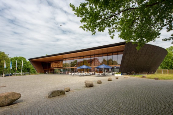Ein modernes, geschwungenes Holzgebäude mit großen Glasfenstern beherbergt ein Café mit Sitzgelegenheiten im Freien; im Vordergrund befinden sich ein mit Steinen gepflasterter Platz und verstreute Felsen.