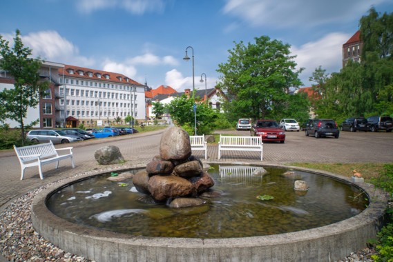 Ein runder Brunnen mit aufgeschichteten Steinen steht in einem gepflasterten Hof, umgeben von Bänken, geparkten Autos, Bäumen und mehrstöckigen Gebäuden unter blauem Himmel.