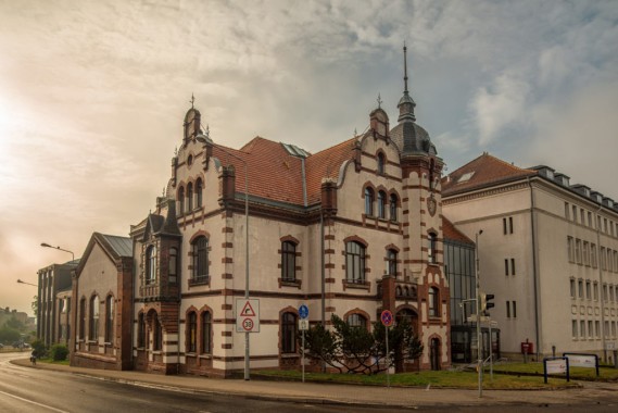 Ein historisches, verziertes Gebäude mit roten Dachziegeln und dekorativem Mauerwerk steht an einer Straßenecke in der Nähe moderner Gebäude unter einem teilweise bewölkten Himmel.