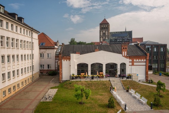 Ein weißes Gebäude mit großen Bogenfenstern und Sitzgelegenheiten im Freien befindet sich zwischen modernen und historischen Strukturen, mit Bänken und einer kleinen Rasenfläche davor. Im Hintergrund ist ein Kirchturm zu sehen.