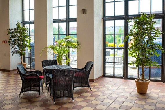 Ein lichtdurchfluteter Innenbereich mit vier schwarzen Korbstühlen um einen Tisch, Topfpflanzen, Fliesenboden und großen Fenstern mit Blick auf eine Außenterrasse.