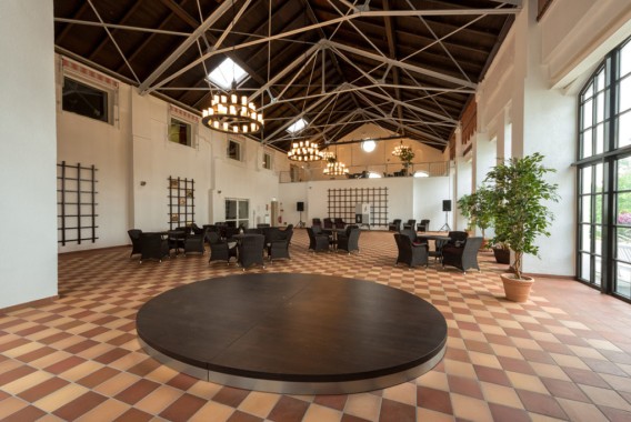 Geräumige Halle mit hoher Decke, Fliesenboden, Korbstühlen um Tische, Topfpflanzen, großen Fenstern und einer runden Bühne im Vordergrund.