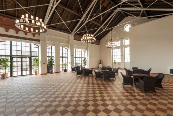 Geräumige Halle mit hohen Decken, großen Fenstern, Fliesenboden, Kronleuchtern, Topfpflanzen und mehreren schwarzen Korbtischen und Stühlen, die auf der rechten Seite angeordnet sind.