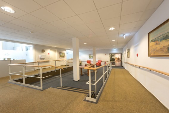 Ein geräumiger Flur mit beigen Wänden und Teppich, Handläufen, einer Rampe, mehreren Stühlen, gerahmten Bildern und heller Deckenbeleuchtung.