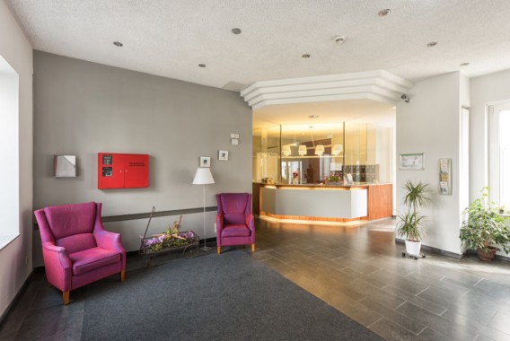 Eine moderne Lobby mit zwei lilafarbenen Sesseln, einer grauen Wand mit einem roten Brandschutzkasten, Topfpflanzen und einem Empfangstresen im Hintergrund.