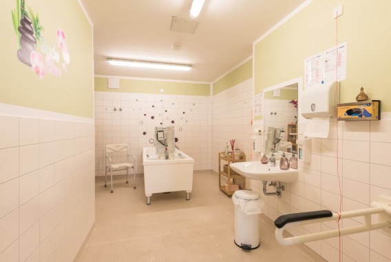 Ein geräumiges, barrierefreies Badezimmer mit Badewanne, wandmontierten Haltegriffen, Stuhl, Waschbecken, Abfalleimer und Notrufkabel, das für Menschen mit Mobilitätsproblemen konzipiert ist.