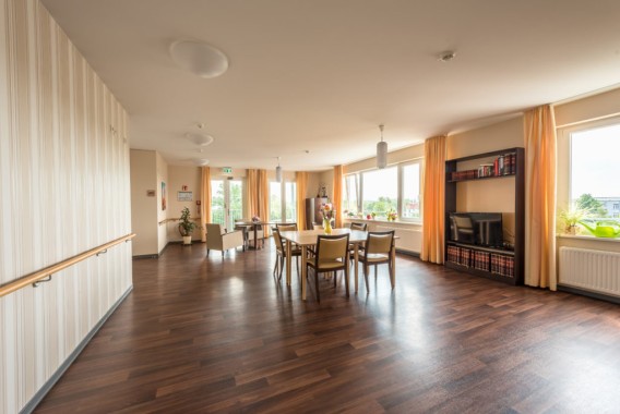 Ein geräumiger, heller Raum mit Holzfußboden, mehreren Stühlen an Tischen, einem Fernseher, Bücherregalen, großen Fenstern und cremefarbenen Vorhängen.