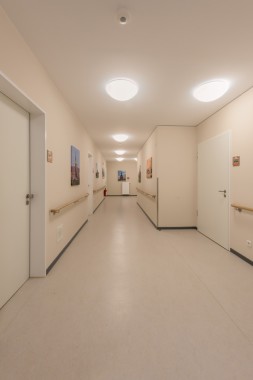 Ein sauberer, leerer Flur mit beigen Wänden, weißen Türen, Handläufen, Deckenleuchten und gerahmten Bildern an den Wänden.