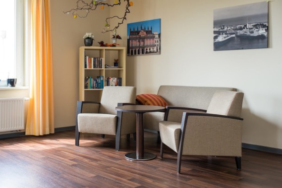 Ein Wohnzimmer mit zwei beigen Sesseln, einem kleinen runden Tisch, einem passenden Sofa, einem Bücherregal, Wandkunst und gelben Vorhängen neben einem Fenster.