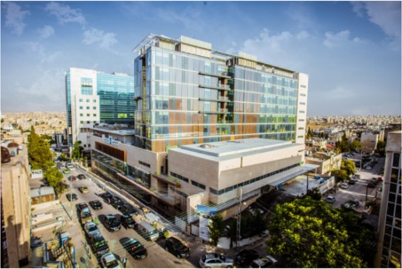 Ein modernes, mehrstöckiges Gebäude aus Glas und Stein steht an einer belebten Straßenkreuzung, umgeben von geparkten Autos und angrenzenden Gebäuden unter einem klaren Himmel.