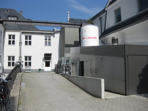 Eine gepflasterte Gasse zwischen weißen Gebäuden mit einem großen Air Liquide-Tank, Gasflaschen und metallverkleideten Versorgungseinrichtungen unter blauem Himmel.