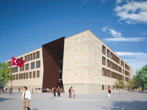 Ein modernes, rechteckiges Steingebäude mit großen Fenstern und einer türkischen Flagge an der Außenseite; mehrere Menschen gehen unter blauem Himmel umher.