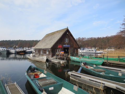 Ein hölzernes Bootshaus mit Strohdach steht am Wasser, umgeben von mehreren kleinen Booten und Yachten, mit Bäumen im Hintergrund.